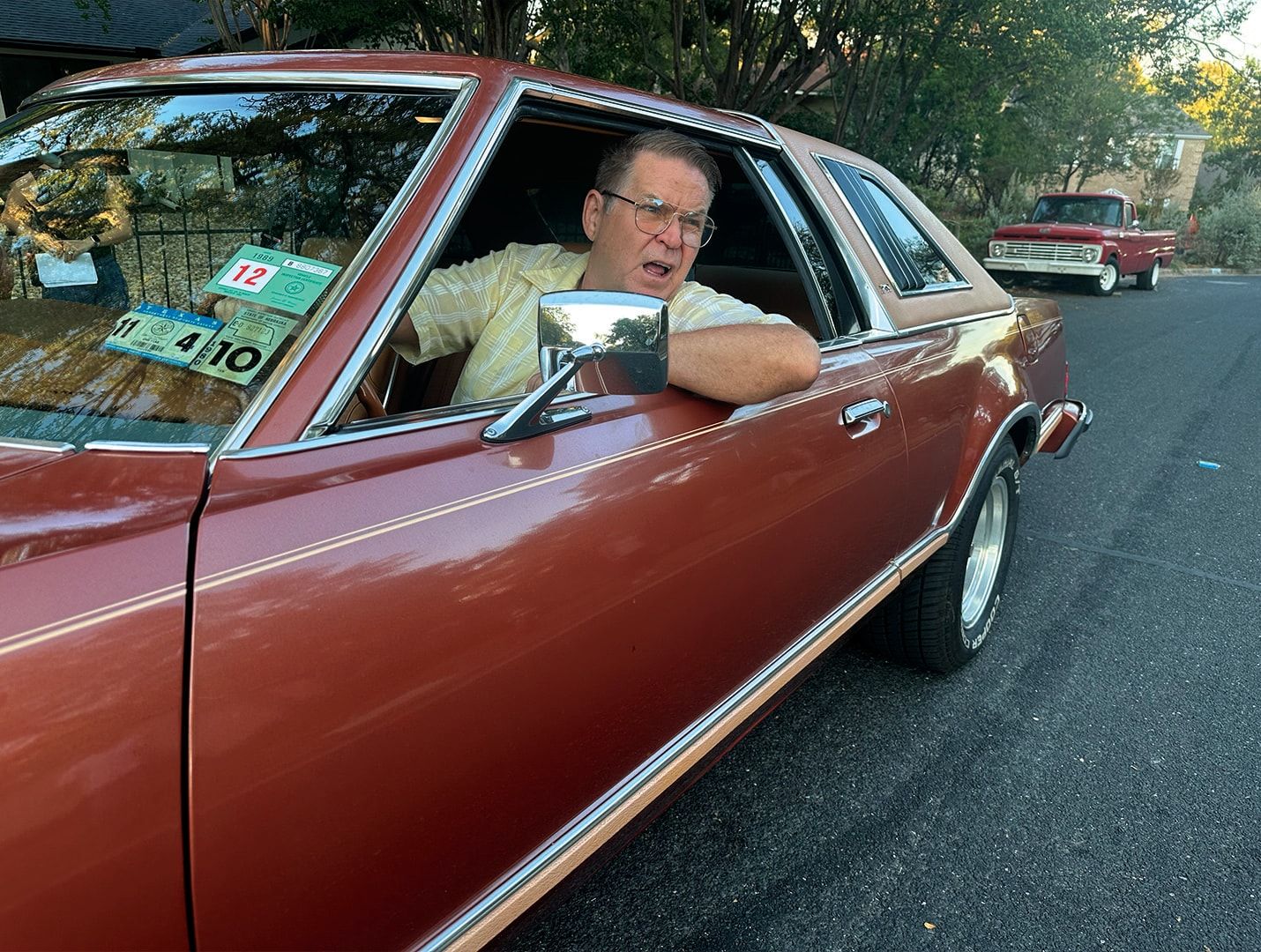 Bill Wise leaning out of the window of a red sedan on a suburban street